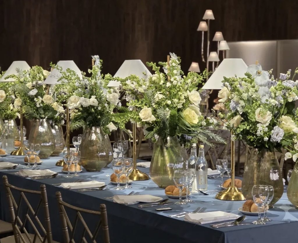 “Elegant event tablescape featuring Baise ribbed glass vases with white and green floral arrangements, gold table lamps, and luxury place settings.”